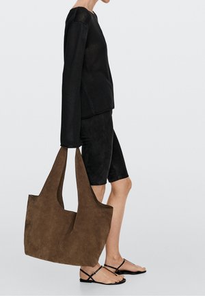 Persoon draagt een zwart lang mouwen topje, zwarte biker shorts en zwarte sandalen en houdt een grote bruine suède totebag vast tegen een effen achtergrond.