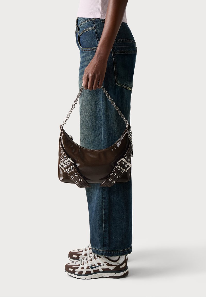 Borsa a mano in pelle verniciata marrone con tracolla in catena argentata, con dettagli decorativi di fibbie e fori. Indossata con jeans a gamba dritta blu.