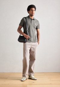 Polo de culoare verde măsliniu cu mâneci scurte, pantaloni bej cu croială largă și geantă crossbody texturată neagră. Adidași cu dungi albe și verzi.