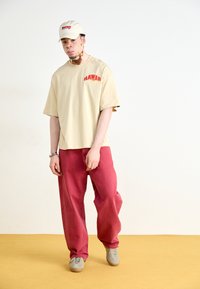 Beige ylisuurustakki, jossa on punainen "HAWAII" -grafiikka, yhdistettyinä väljiin punaisiin housuihin ja harmaisiin lenkkareihin. Vaaleanvärinen lippis viimeistelee asun.