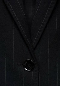 Černý blazer s vertikálními bílými pruhy, s jedním černým knoflíkem a klopovým límcem, vyrobený z texturované látky.
