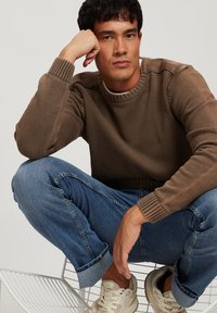 Brązowy sweter z dzianiny z prążkowanymi mankietami i dekoltem, w zestawieniu z podwiniętymi niebieskimi dżinsami i jasnymi sneakersami na metalowym krześle.