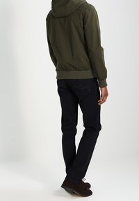 Muž v olivově zelené bundě s kapucí, tmavě modrých džínách a tmavě hnědých botách, stojící s mírně otočeným zády k jednoduchému pozadí.