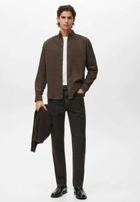 Camisa marrón de botones sobre una camiseta blanca, combinada con jeans oscuros y botas negras. Un suéter de punto está colgado sobre un brazo.