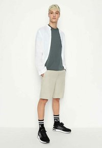 Camicia button-up bianca in lino, t-shirt verde scuro, pantaloni corti beige, sneakers nere e calze a righe. Outfit casual con diverse textures.