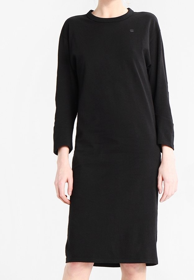 Femme portant une robe noire simple à manches longues, longueur genou, avec un col rond, sur un fond blanc uni.