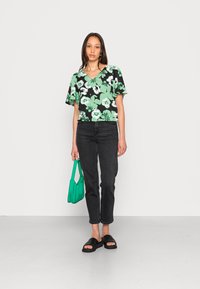 Top floral preto com detalhes verdes, mangas curtas, decote em v; combinado com jeans pretos e sandálias de slide pretas; segurando uma bolsa teal.