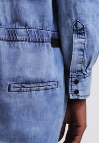 Veste en denim bleu avec une texture légèrement froissée, dotée d'un poignet à boutons et d'une poche subtile avec un bord surpiqué.