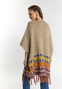 Poncho tricotat în bej, cu modele geometrice colorate la margine. Are margini franjurate și un design lejer, fluid.