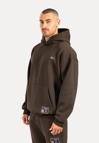 Brauner Kapuzenpullover aus strukturiertem Stoff, mit einer Vorderjacke und einem kleinen Logo. Kombiniert mit passenden braunen Hosen und Etiketten.