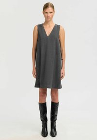 Graues ärmelloses Kleid mit V-Ausschnitt und A-Linien-Form. Hergestellt aus einem strukturierten Stoff, reicht es bis oberhalb des Knies und wird mit hohen schwarzen Stiefeln kombiniert.