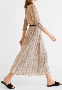 Kevyt, pitkä beige mekko, jossa on teksturoitu käärmeennahkakuosi, kolmen neljänneksen hihat, kireä vyötärö mustalla vyöllä, yhdistetty mustien liukusandaalien kanssa.