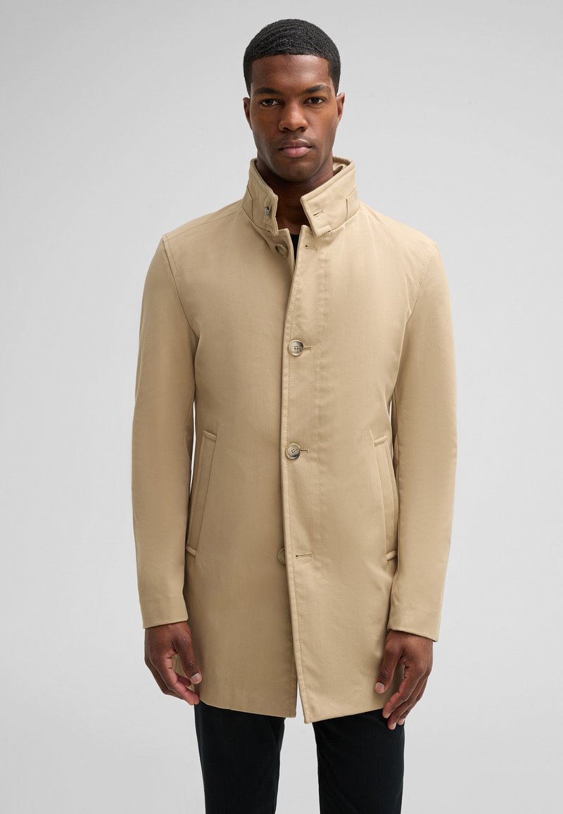 Beige jas met hoge kraag, voorknopsluiting en twee zijzakken; soepele textuur en getailleerde pasvorm. Draagt met zwarte broeken.