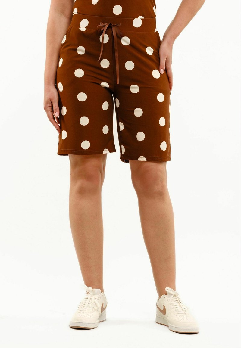 Personne portant un short marron mi-long à pois blancs et des baskets blanches, debout devant un fond uni.