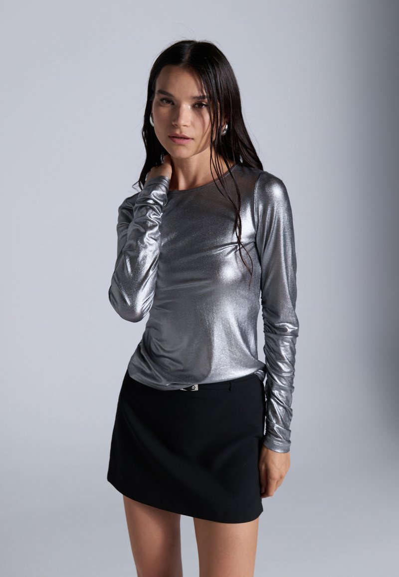 Sidabrinė metalinė ilgomis rankovėmis bluzonė su prigludusia forma, derinama su juoda mini sijonu. Lygi tekstūra ir elegantiška silueto forma pabrėžia aprangą.