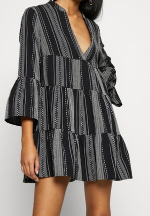 Femme portant une robe noire à rayures verticales blanches et manches évasées, avec un profond décolleté en V et une jupe à étages.