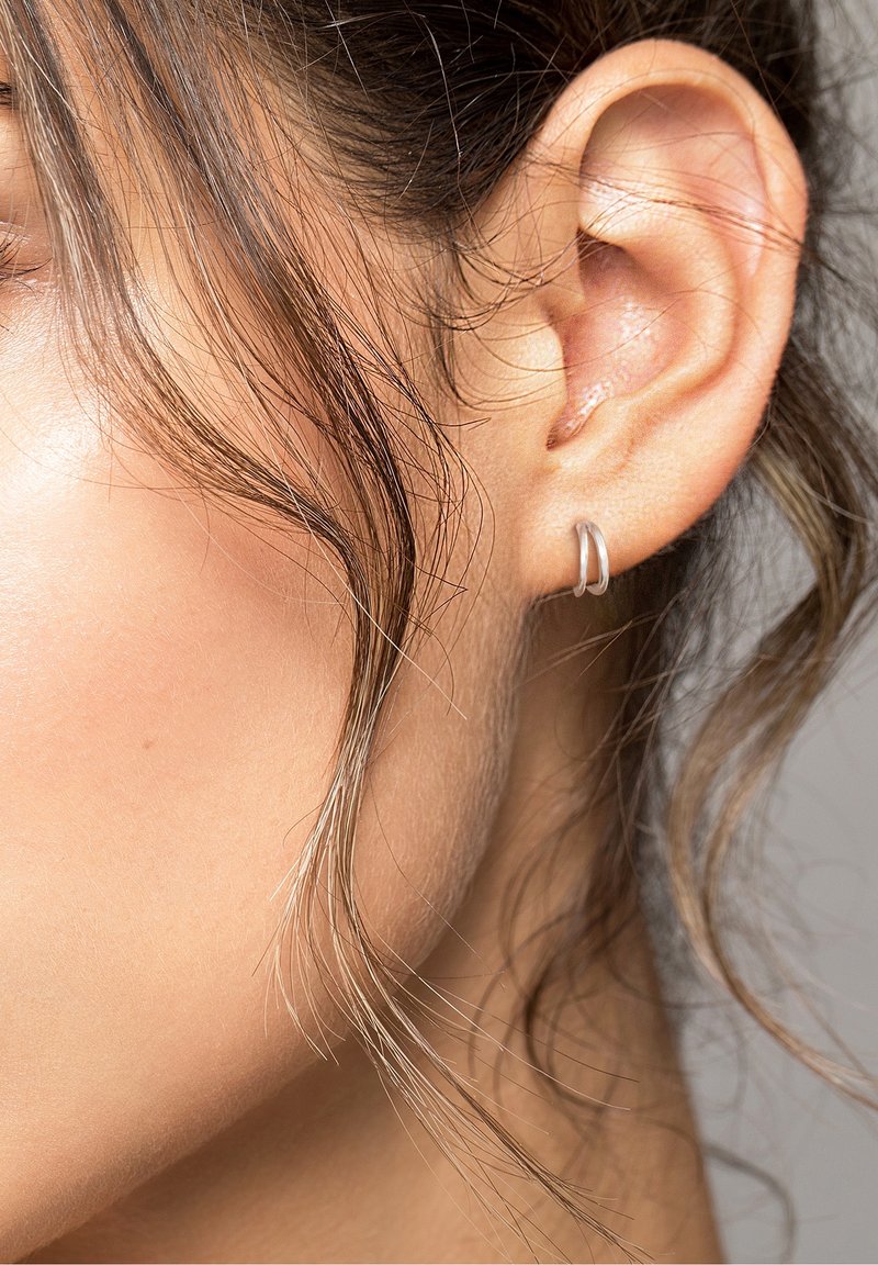 Gros plan sur l'oreille d'une femme avec une petite boucle d'oreille argentée et des mèches brunes lâches encadrant son visage.