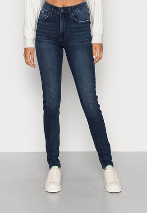 Frau trägt dunkelblaue Slim-Fit-Jeans, weiße Sneakers und einen weißen, kurz geschnittenen Pullover, steht vor einem einfarbigen, hellen Hintergrund.