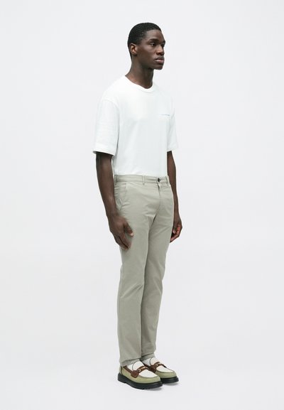 Λευκό κοντομάνικο μπλουζάκι με ανοιχτό μπλε κ είμενο, συνδυασμένο με ανοιχτό γκρι παντελόνι slim-fit και μπεζ παπούτσια με καφέ λεπτομέρειες.