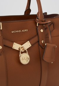 Sac à main Michael Kors en cuir marron texturé avec plaque logo dorée, détail cadenas doré et étiquette en cuir assortie suspendue à la poignée.