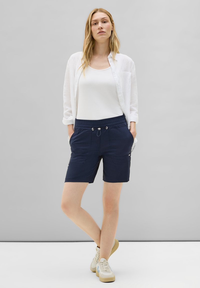 Shorts bleu marine en tissu léger avec une taille élastique, un cordon de serrage et des poches latérales. Associés à un débardeur blanc et à une chemise.