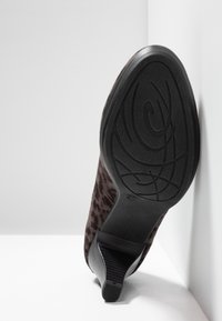 Chaussure pour femme à talon noir avec dessus à imprimé léopard, semelle montrant le motif de la bande de roulement, reposant contre un mur blanc à angle sur un sol blanc.