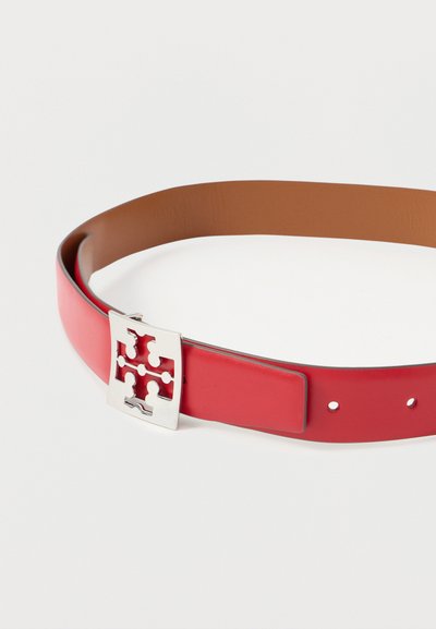 Κόκκινη δερμάτινη ζώνη με καφέ εσωτερική πλευρά και ασημένια τετράγωνη αγκράφα με συμμετρικό σχέδιο κοπής, τοποθετημένη σε λευκό φόντο.