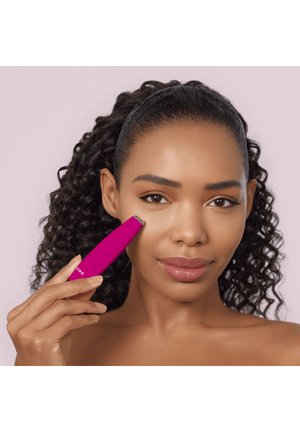Femme aux cheveux bouclés utilise un petit appareil rose pour les soins de la peau sur sa joue droite sur un fond rose clair uni.