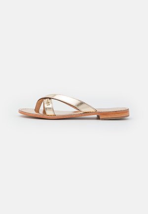 Flad sandal med metallisk guld krydsede læderstropper og en tan lædersål med lav træsål, vist fra siden mod en ensfarvet baggrund.