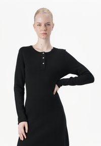 Black long-sleeve dress made of knit fabric, featuring a round neckline and three white buttons down the front, with a fitted silhouette.