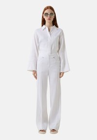 Witte linnen blouse met lange klokmouwen, gecombineerd met hoge witte broeken. Model draagt ronde zonnebril en platte sandalen.