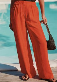 Pantalons larges, orange vif avec un design texturé et plissé. Associés à un sac à main en osier noir et des sandales noires.