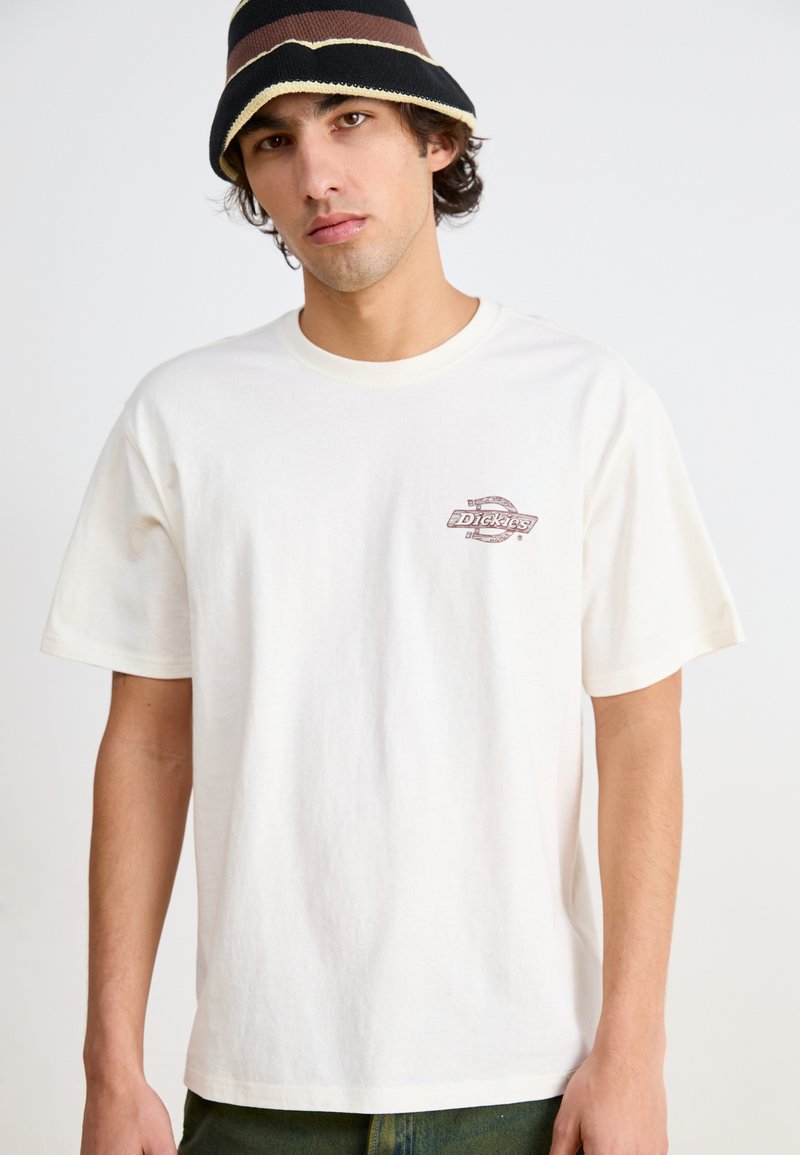 T-shirt in cotone crema con maniche corte, scollatura tonda e un piccolo logo grafico marrone sul lato sinistro del petto. Taglio semplice e rilassato.