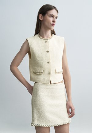 Ženska v brezrokavnem kremnem tweed jopiču s školjčnimi gumbi in ujemajočo se krilo z okrasnim obrobom iz školjk, stoji ob enobarvnem sivem ozadju.