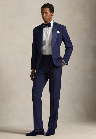 Άνδρας που φοράει σκούρο μπλε σμόκιν με σατέν πέτα, λευκό πουκάμισο με πιέτες, μαύρη παπιγιόν και βελούδινα μπλε μοκασίνια, στέκεται με το ένα χέρι στην τσέπη.