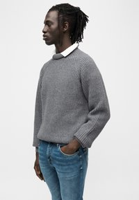 Γκρι πλεκτό πουλόβερ με ριμπ υφή, στρογγυλό γιακά και ραγκλάν μανίκια, συνδυασμένο με μπλε τζιν. Απλός και χαλαρός σχεδιασμός.