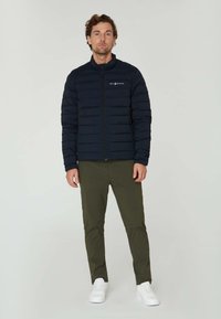 Marinblå pufferjacka med hög krage, horisontell quiltad design och dragkedja framtill. Kombinerad med olivgröna byxor och vita sneakers.