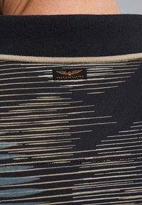 Chemise noire avec un col beige étroit et une étiquette. Présente des lignes horizontales de différentes couleurs, y compris bleu clair et crème.