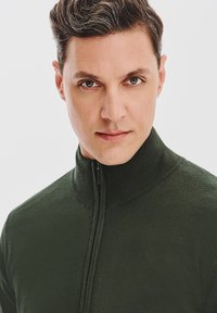 Ciemnozielony sweter na zamek z wysokim kołnierzem. Miękki, fakturowany materiał, z zamkiem z przodu i prążkowanym kołnierzem dla dodatkowego efektu.