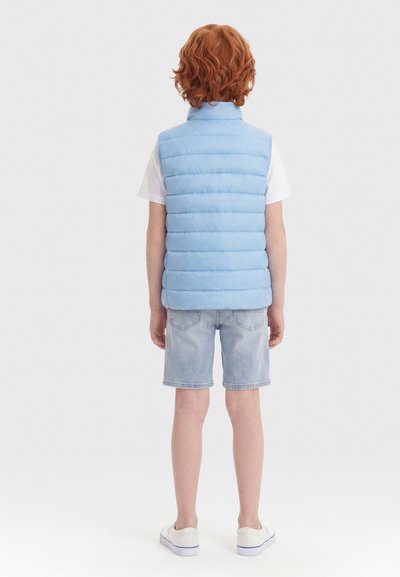 Παιδί με κόκκινα μαλλιά που φοράει ανοιχτό μπλε φουσκωτό γιλέκο, λευκό κοντομάνικο πουκάμισο, τζιν σορτς και λευκά αθλητικά, στέκεται με την πλάτη προς το μέρος μας.