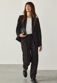 Černé sako přes bílý žebrovaný top, v kombinaci s černými elegantními kalhotami a černými koženými botami. Drží malou černou kabelku.