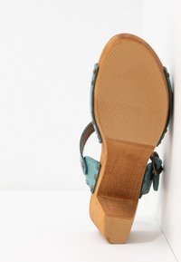 Sandale avec un talon épais en bois et une semelle texturée, présentant des brides turquoise avec des clous en métal et une boucle, vue de dessous.