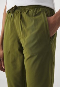 Olivově zelené sportovní kalhoty s elastickým pasem, šňůrkou a bočními kapsami. Hladká textura s jemnými švy.