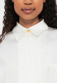 Blusa branca com colarinho estruturado, apresentando pregas e um botão dourado no pescoço. Textura suave, design minimalista.