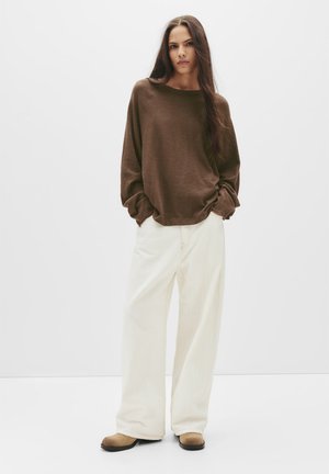 Bruine lange mouw top met een ontspannen pasvorm, gecombineerd met crèmekleurige wijde broek en beige schoenen. Soepele textuur, minimalistisch ontwerp.