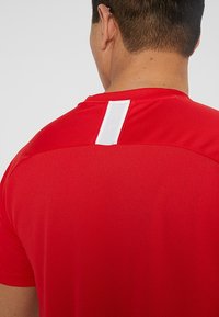 Camiseta deportiva roja con una tela texturizada, con una etiqueta blanca y detalles de malla en la parte superior de la espalda, escote redondeado y mangas cortas.
