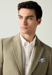 Khakifarbener Blazer mit strukturiertem Finish, weißes Hemd mit Knopfleiste und ein floral bedrucktes Einstecktuch mit blauen und rosa Mustern.