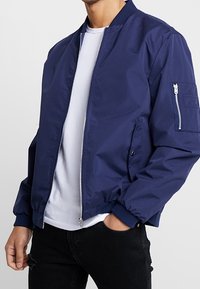 Námořnicky modrá bomber bunda z hladké látky, s předním zipem, žebrovanými manžetami a boční kapsou na zip. Nošená přes bílé tričko.