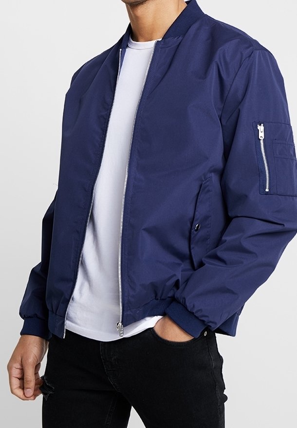 Námořnicky modrá bomber bunda z hladké látky, s předním zipem, žebrovanými manžetami a boční kapsou na zip. Nošená přes bílé tričko.