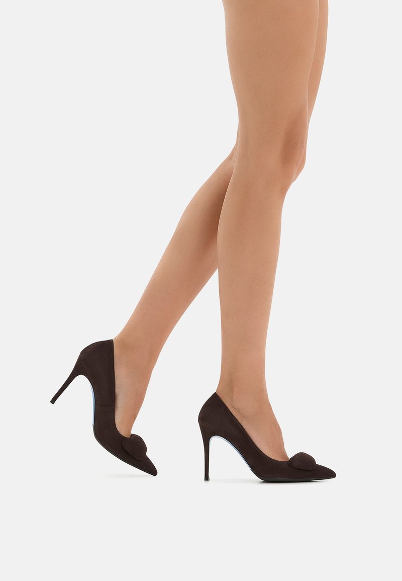 Pompe in suede marrone con tacco alto, punta affusolata, tagli laterali curvi e un accentuazione con pompon sulla punta. Presentano un tacco sottile e una texture liscia.